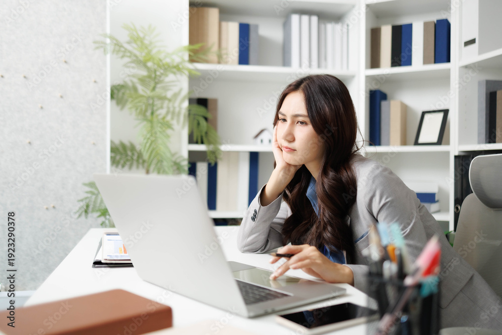 Fototapeta premium A woman is sitting at a desk with a stack of papers in front of her. She is looking at a piece of paper and she is in a state of confusion or frustration 