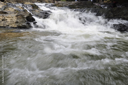 Fast flowing waterfall river rushing over dark rocks in lush green forest. The water appears white and foamy  cascades uneven stones creating small rapids. 