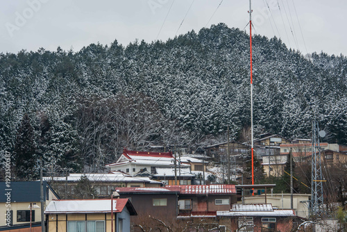 Traditional Japanese houses nestled on a snowy hillside with a tall red and white communication antenna and evergreen forest.