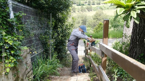 Handyman wearing a face mask and overalls working on outdoor home renovation, diligently sanding a wooden fence post with an electric power tool for maintenance
