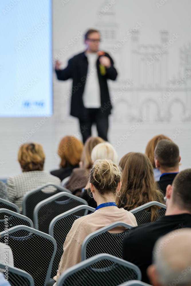 custom made wallpaper toronto digitalAudience listening to presenter at business conference, focused attendee with lanyard in foreground, blurred speaker on stage, professional event atmosphere.