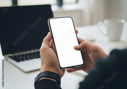 A person holding a smartphone with a blank screen in front of a laptop on a white desk in a modern office space