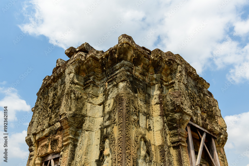 Fototapeta premium A weathered sandstone tower adorned with intricate carvings rises against a backdrop of blue sky and scattered clouds at Phnom Bakheng temple near Siem Reap, Cambodia. The historic structure displays