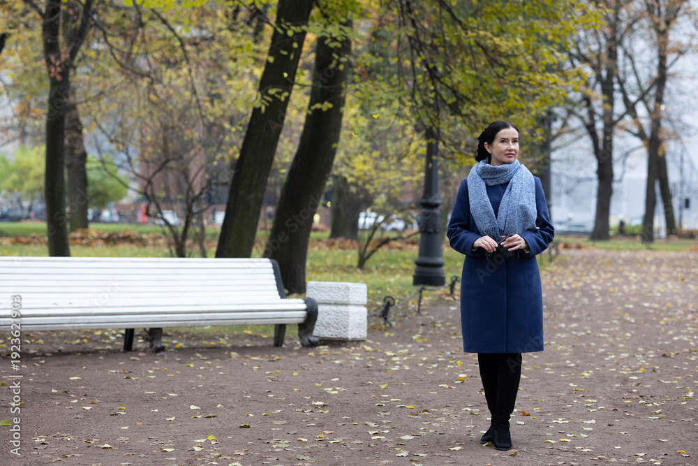Fototapeta premium Mature woman walking on city sidewalk during autumn near residential architecture.