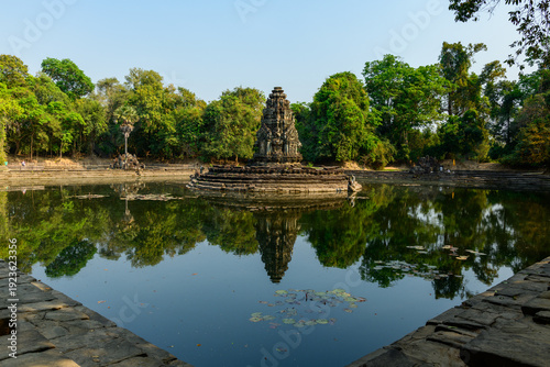 фотографія Stone shrine rises from the center of a tranquil pond at Neak Pean temple, surrounded by lush tropical trees and calm water in Cambodia's Angkor region