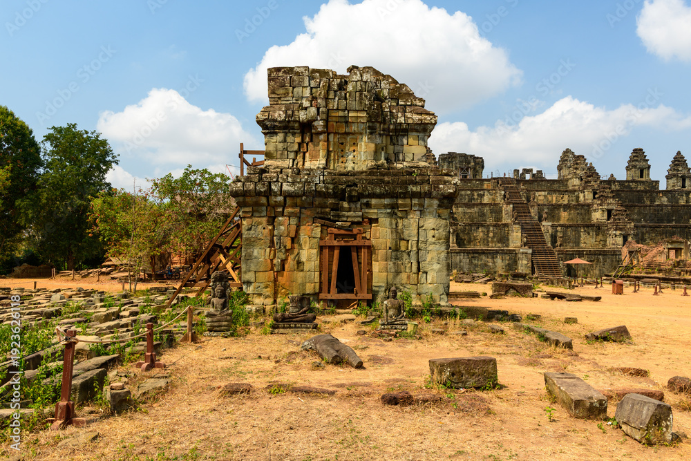 Fototapeta premium Weathered stone structures and scattered statues stand on the sunlit grounds of Phnom Bakheng temple near Siem Reap, Cambodia. The historic site features layered masonry, dry earth, and a backdrop of