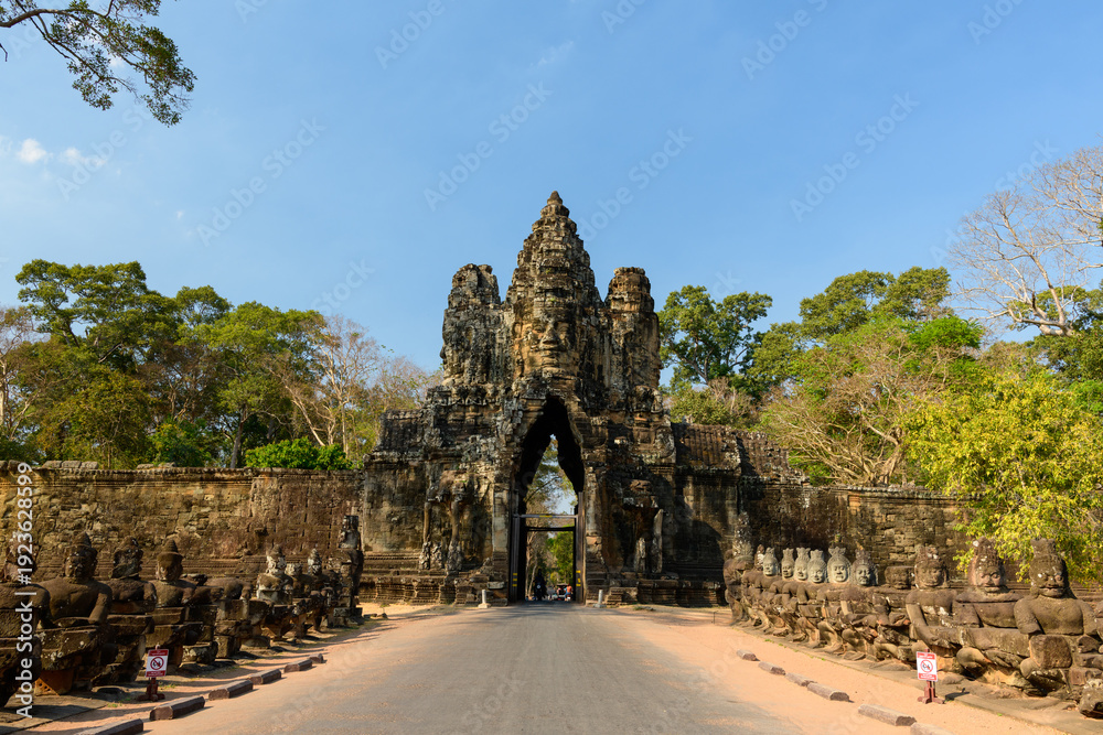 Fototapeta premium Wide frontal view of the iconic south gate of Angkor Thom near Siem Reap, Cambodia, featuring towering carved stone architecture flanked by rows of guardian statues. The ancient Khmer gateway stands