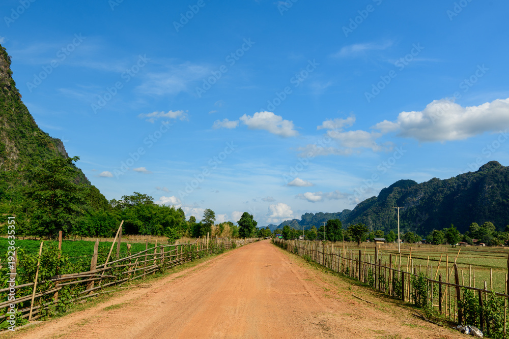 Fototapeta premium A straight red earth road runs through open farmland bordered by rustic wooden fences, with green fields and distant limestone hills beneath a vivid blue sky. The landscape features natural textures
