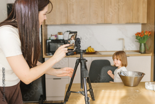 Mother recording daughter cooking on smartphone in kitchen, family creating home video content and baking together