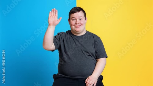Happy young man with Down syndrome waving his hand, smiling at the camera on a blue and yellow background.
