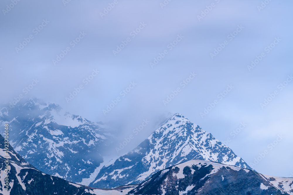 Obraz premium Mont Lachat's pointed summit emerges through drifting mist, with streaks of melting snow on rugged slopes. Soft blue light and overcast sky create a tranquil, ethereal alpine mood.