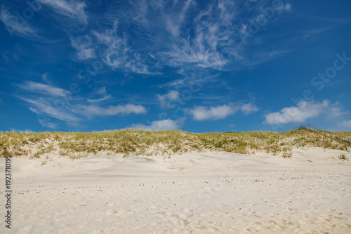 Scenic coastal landscape featuring a sandy beach with dry grass dunes and a wooden staircase leading to the sea under a bright blue sky with wispy cirrus clouds for travel and nature concepts