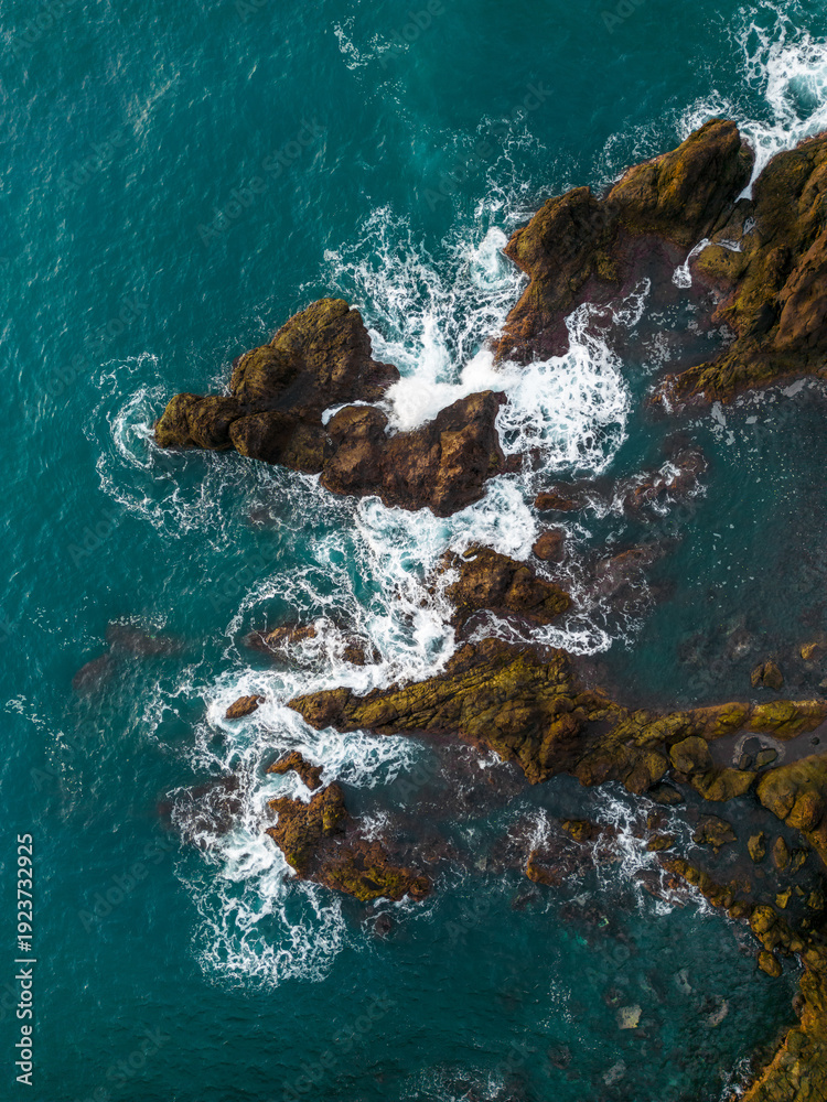 Obraz premium Drone view of powerful Atlantic Ocean waves crashing against rugged volcanic rocks along the dramatic coastline of Madeira Island, Portugal.