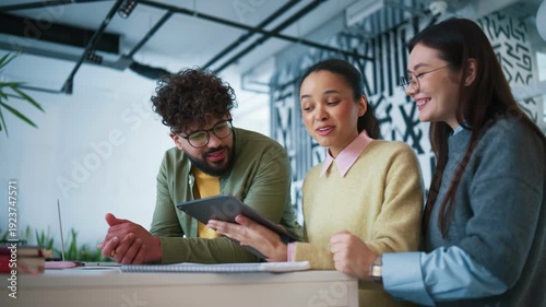 Modern interior workspace. Colleagues gathering. Team reviewing tablet and identifying issue. Smiling coworkers discussing solution and coordinating next steps during collaborative work session.