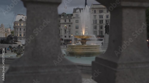 Artistic static shot of an illuminated fountain and the General Napier statue in Trafalgar Square, framed through stone railings at twilight.