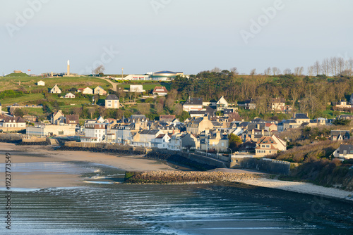 Charming coastal village with rows of traditional houses nestled against lush green hills in Arromanches-les-Bains, Normandy. Soft evening sunlight highlights the peaceful shoreline and tranquil