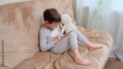 Child boy sits on couch, playing with a digital tablet next to his soft toy. Modern technology for kids entertainment and education concept.