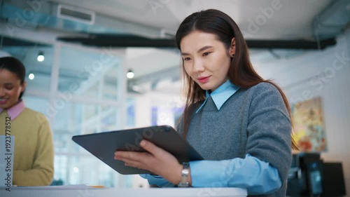 Focused office portrait at work desk. Asian woman holding tablet computer and reading digital information. Female professional checking online report while colleague smiling in background.