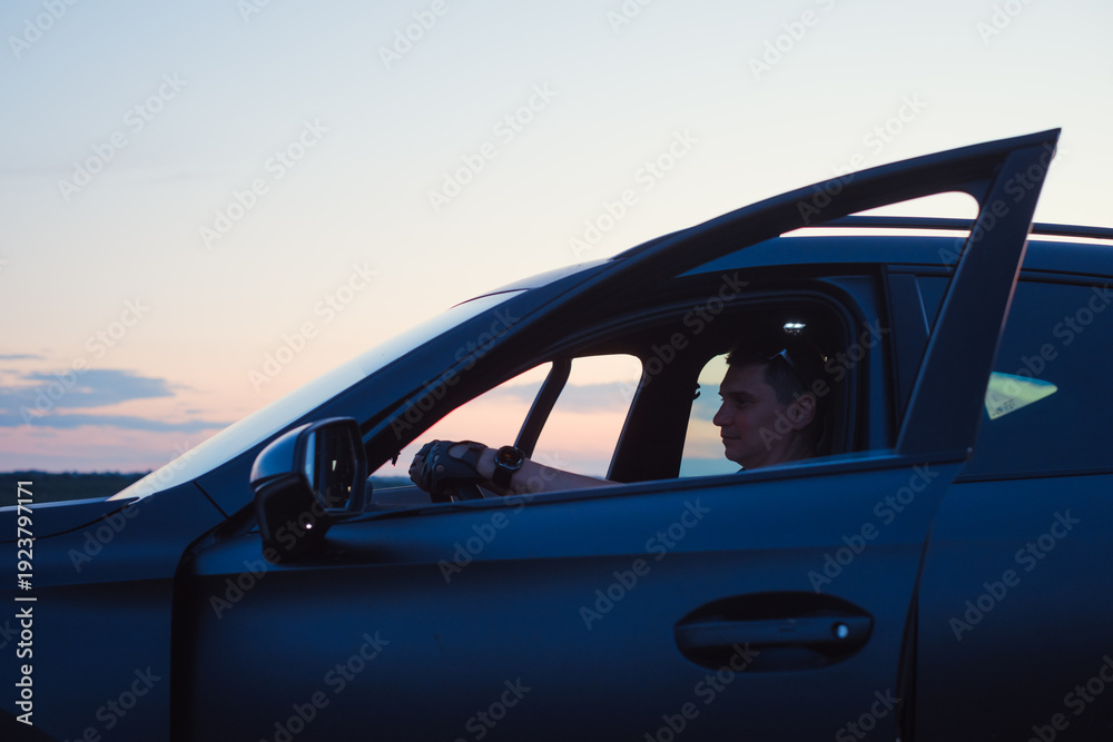 Naklejka premium Side view of male driver sitting in modern dark car with opened door holding steering wheel. Evening road trip concept with beautiful sunset sky background.