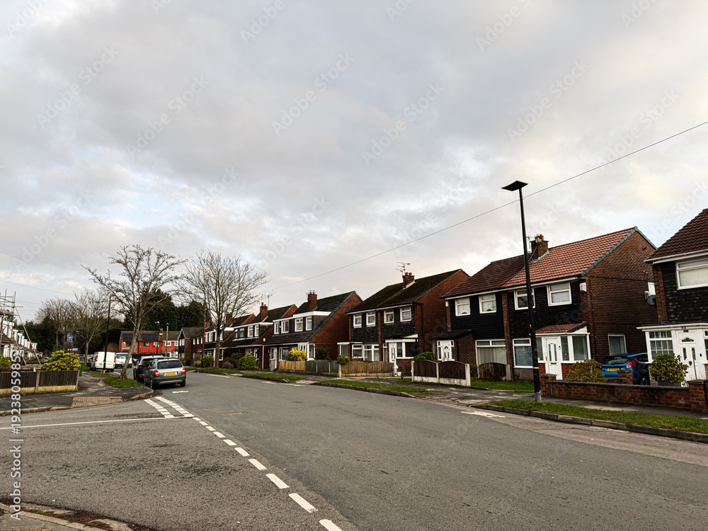 Obraz premium Residential street in Manchester UK with row houses, parked cars, and a cloudy sky