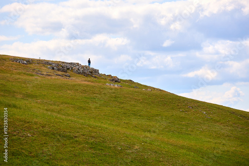 Konstfotografi Male standing on scenic hilltop with expansive green grass and cloudy blue sky