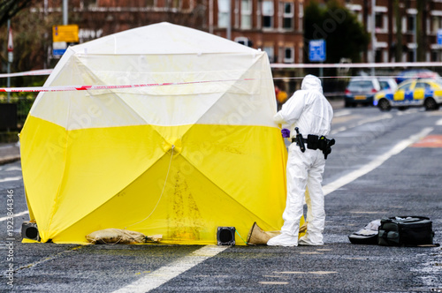 A scene of crime officer wearing a white boiler suit looks inside an examination tent at a crime scene