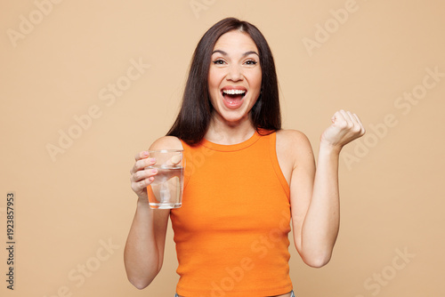 Fotografi Young woman wear orange casual clothes hold in hand glass drink water do winner gesture isolated on plain pastel light beige background