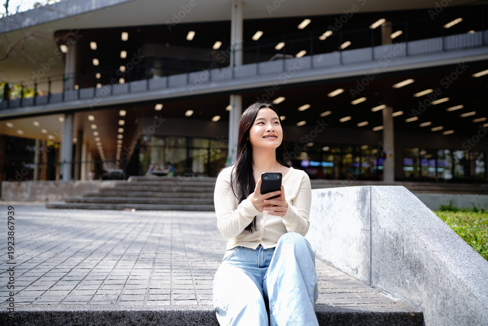 Fototapeta premium Asian student relaxing on campus stairs using smartphone