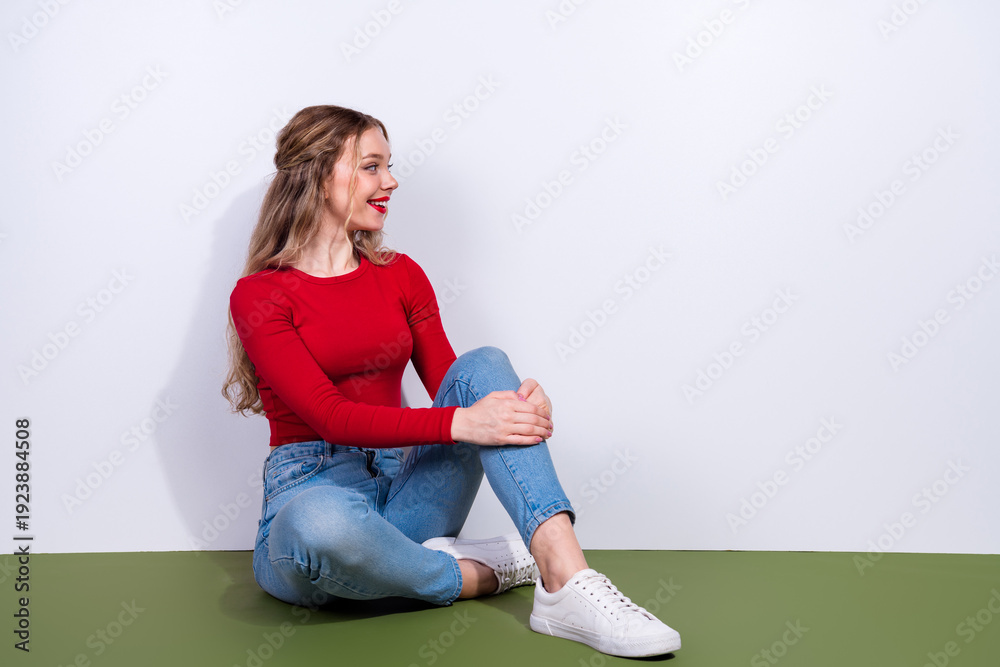 Naklejka premium Young woman in red top sits on green floor smiling with crossed legs in casual fashion lifestyle portrait
