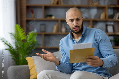 Fotografie Man sitting on a couch at home, opening a received letter and looking confused o