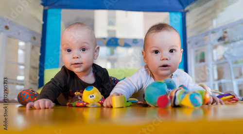 Obraz na płótnie Two adorable twins are on their bellies in a lively playroom, surrounded by an array of colorful toys