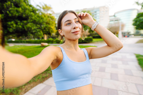 Free Download Selfie time! A happy woman takes a selfie after a workout in a sunny park. A fit woman blogs, videoconferences, and enjoys the sunny day. Concepts of sports, recreation, and blogging. - GFXLoad