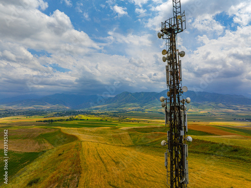 Telecommunications tower stands tall in expansive rural landscape. Parabolic dishes and antennas dot the structure against a backdrop of rolling hills and mountains under a partly cloudy sky.