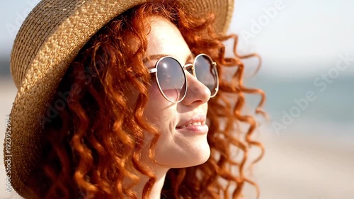 Happy woman with vibrant red curly hair enjoys sunshine wearing stylish sunglasses outdoors