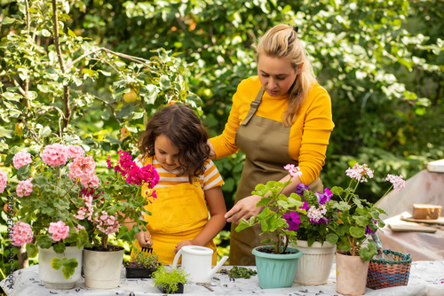 A woman mom and a kid girl child carefully tend to vibrant potted flowers in a lush garden, wearing gardening gloves and aprons, surrounded by greenery and blooming plants.