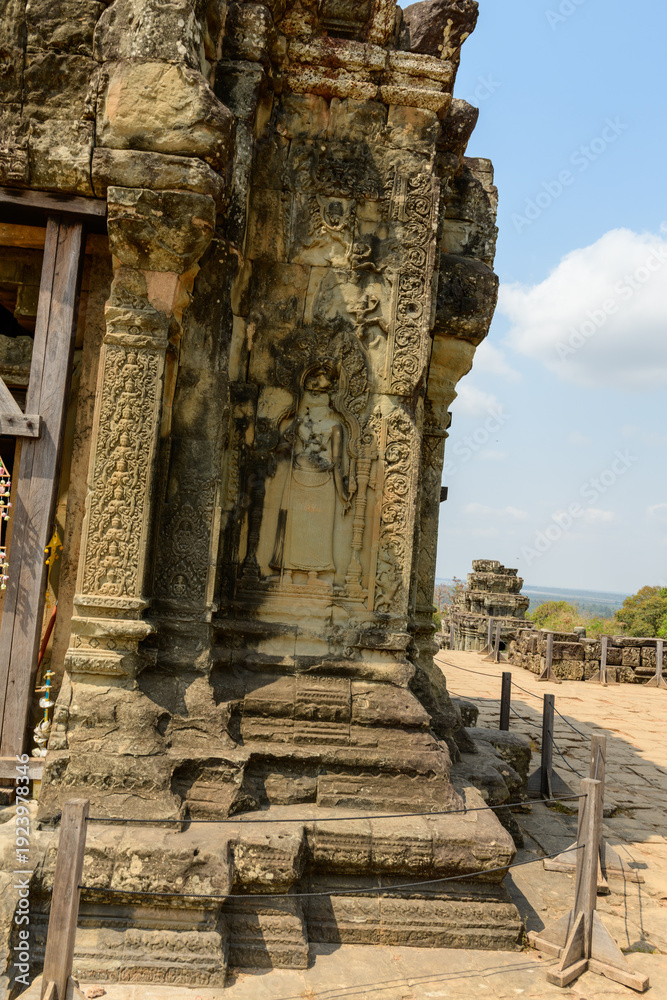 Fototapeta premium Intricate bas-relief of a standing figure adorns the weathered sandstone wall of Phnom Bakheng temple near Siem Reap, Cambodia. The historic structure features ornate patterns and ancient motifs