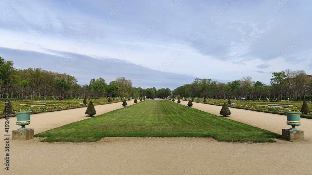 Obraz premium There are many flower beds in the park next to dirt paths and grassy lawns. Tulips, daffodils and other flowers grow on them. Various trees grow nearby. Sunny spring weather and cloudy sky
