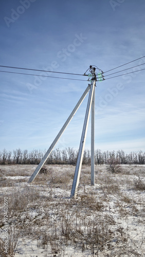 A tall metal power pole stands in a snowy field under a clear blue sky. Sparse vegetation surrounds the area, indicating a cold climate.