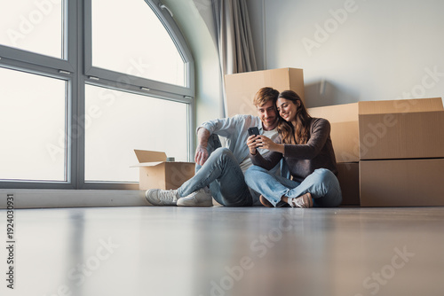 A young couple sits on the floor of their new apartment surrounded by moving boxes, looking out the window while imagining future memories and planning a comfortable modern home together