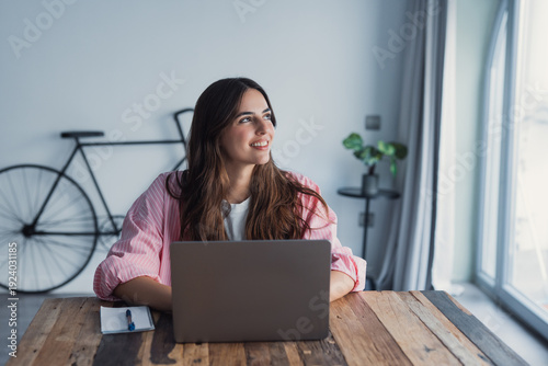 Принти на полотні A dreamy young woman sits at her desk with a laptop, gazing into the distance wh