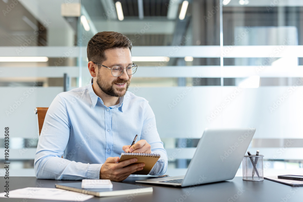 Obraz premium Young businessman wearing glasses sitting at a desk, looking at his laptop screen and writing diligent notes in a spiral notebook, showcasing productivity in a modern office environment