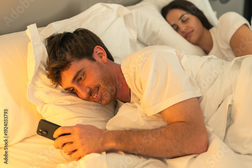 Valokuva A disloyal partner uses his smartphone under the covers while his brunette girlf