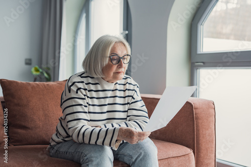 Fotografie Focused elderly 60s woman sit on sofa in living room reading postal corresponden