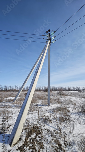 Electricity poles stand in a snowy landscape under a clear blue sky. The ground is covered with snow, and trees are visible in the background.