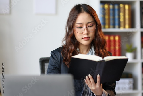 Asian female lawyer reading a law book and studying a case sitting at her desk in a law firm or legal office, she is wearing glasses and turning a page of the book.