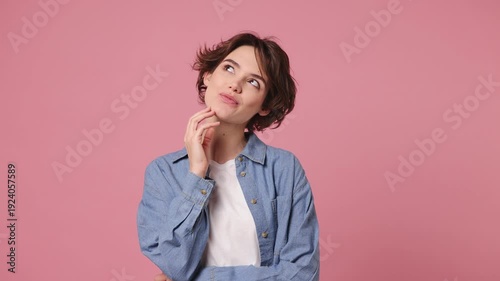 Young dreamful fun woman wears blue shirt white t-shirt casual clothes look around think dream put hand prop up on chin lost in thought and conjectures isolated on plain pastel light pink background