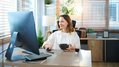 woman sitting at home office table has breakfast with desktop pc. Smiling relaxed young lady hybrid working on computer in modern house