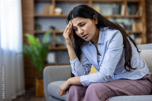Φωτογραφία Young woman with dark hair sitting on a sofa at home, holding her forehead and w