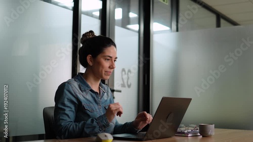Young business woman in denim shirt smiling while closing laptop at end of workday in contemporary corporate office space