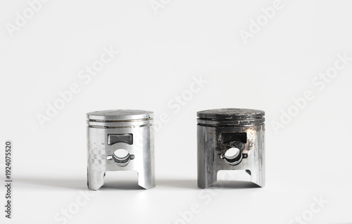 The photograph shows two metal pistons for an internal combustion engine against a light background. On the left is a new, clean piston. On the right is an old one, covered in dark carbon deposits and